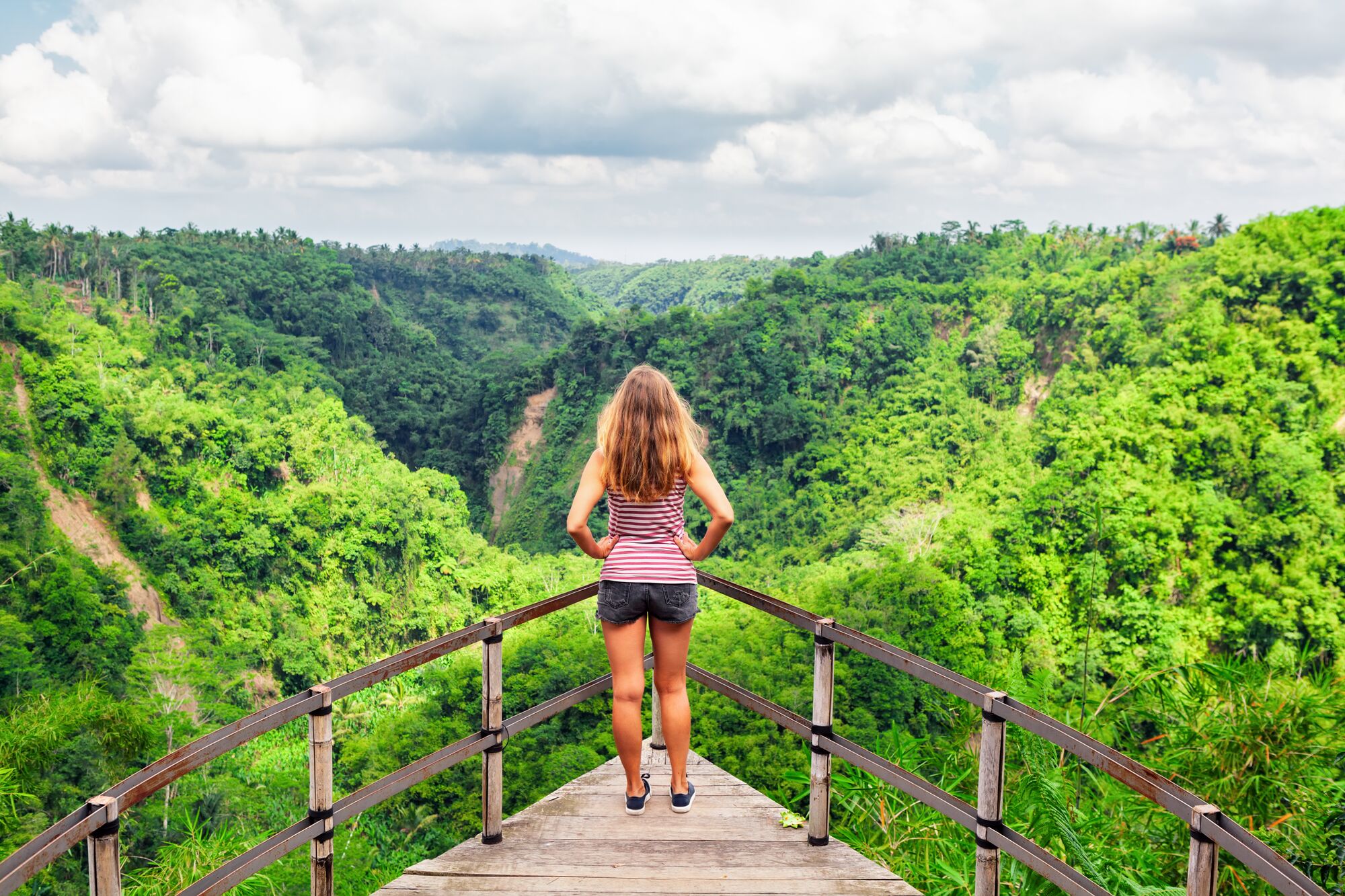 A woman stands on a wooden platform, overlooking a lush green valley in a tropical landscape, embodying adventure and nature's beauty.