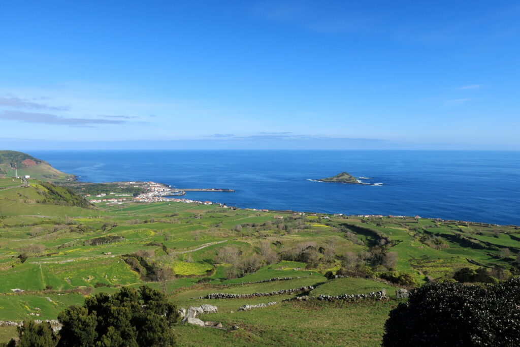 Panoramic view of green hills leading to a coastal village and blue ocean, with a small island visible in the distance under a clear sky.