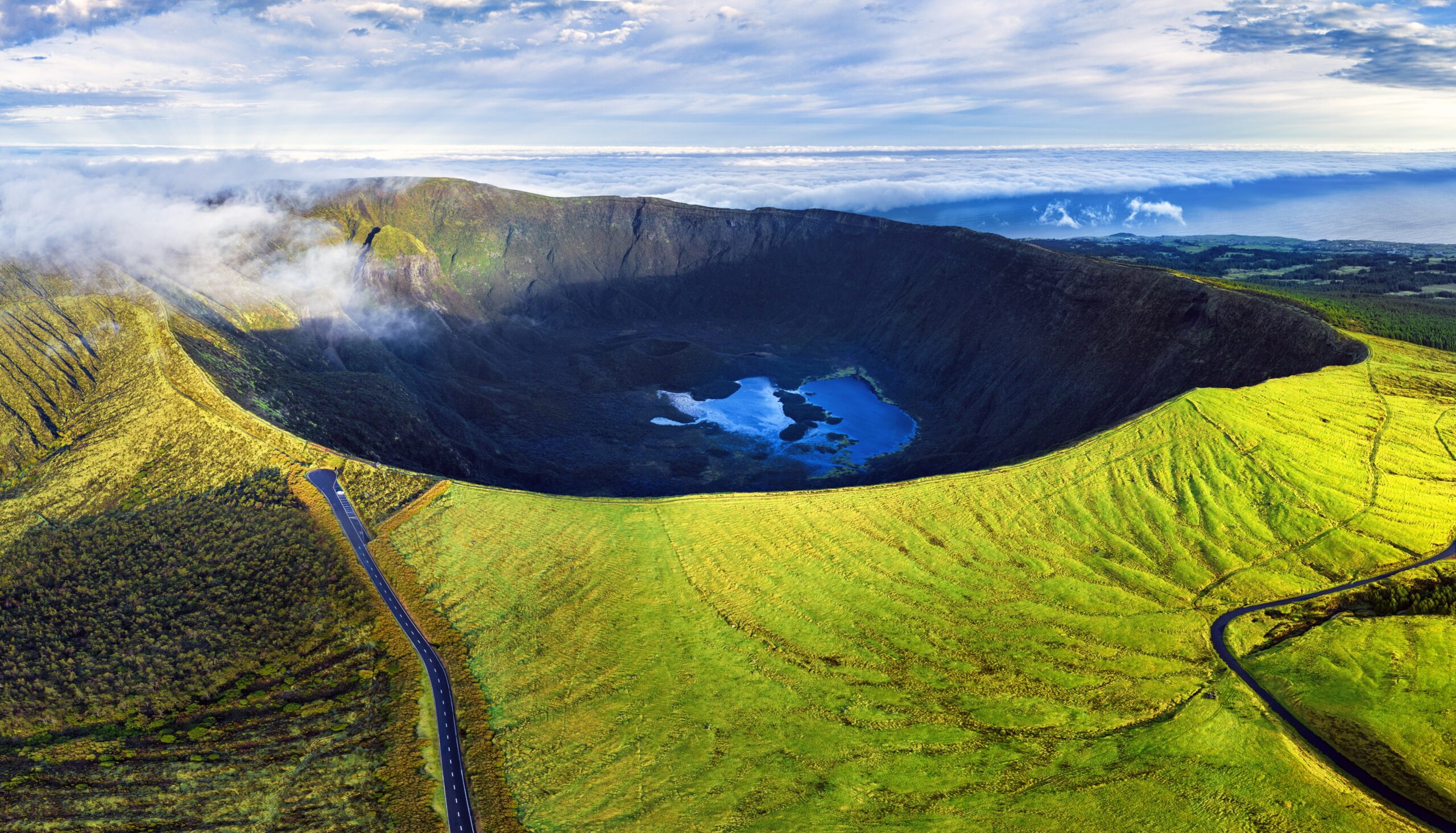 Aerial view of a volcanic caldera with a blue lake at its center, surrounded by lush green hills and winding roads under a cloudy sky.