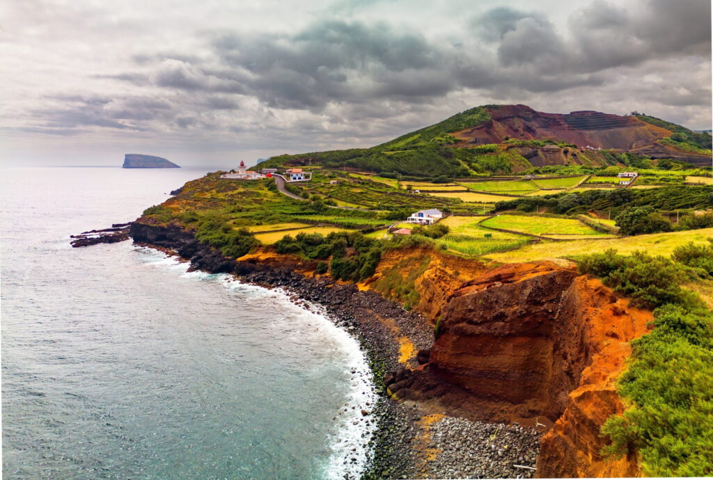 Coastal landscape featuring steep cliffs and lush fields, with a lighthouse and houses overlooking the ocean under a cloudy sky.